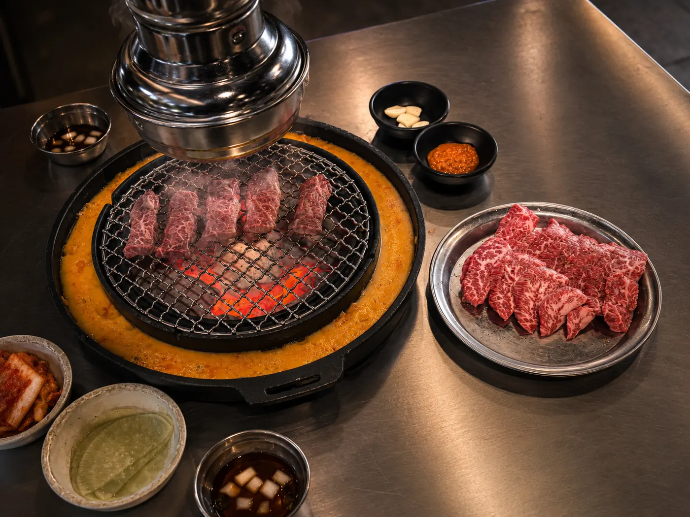Marbled beef grilling over charcoal on a tabletop grill with a golden cheese ring, side dishes, and a plate of raw beef